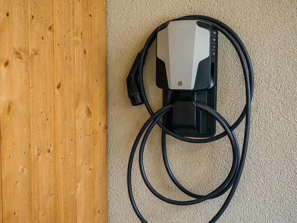 An EV charger installation is mounted on a stucco wall beside a wooden panel, with its charging cable coiled neatly.