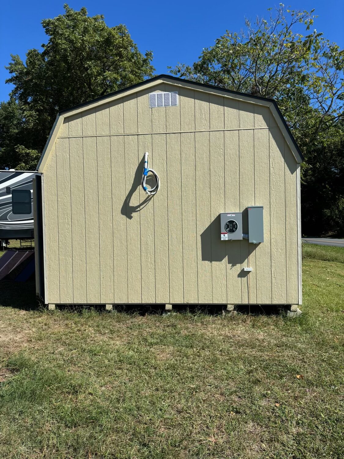 A beige utility shed sits on a grassy lawn, with an electrician-installed electric meter box and coiled cable mounted on its side. Trees and part of a camper are in the background.