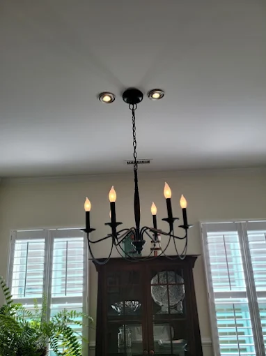 A black metal chandelier with six lit bulbs hangs from the ceiling above a wooden cabinet, in front of two windows with white shutters and plants visible below.
