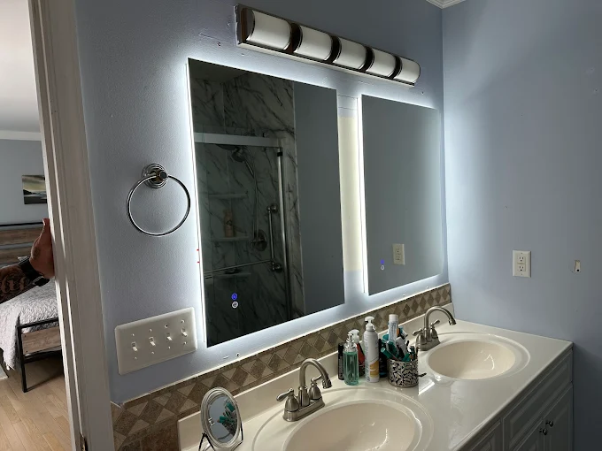 A bathroom with a double sink vanity, various toiletries, a lit LED mirror, towel ring, and a wall-mounted light fixture above the mirror.