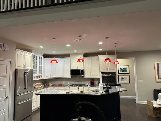 Modern kitchen with white cabinets, stainless steel appliances, and a large island. Four red pendant lights hang above the island. Various kitchen items are on the counters.
