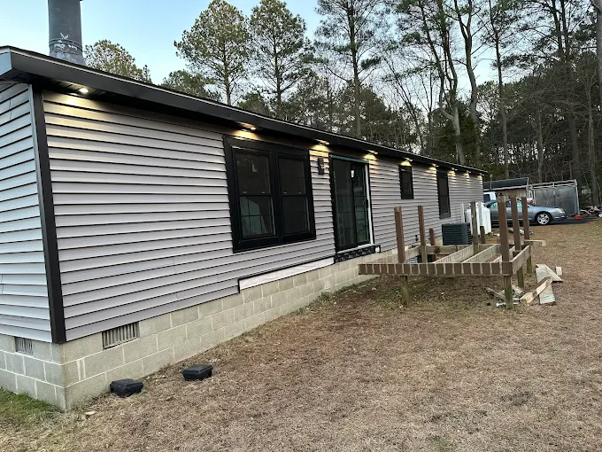 A single-story house with gray siding, black trim, and outdoor lights. An unfinished wooden deck is attached. Trees and a metal trailer are in the background.