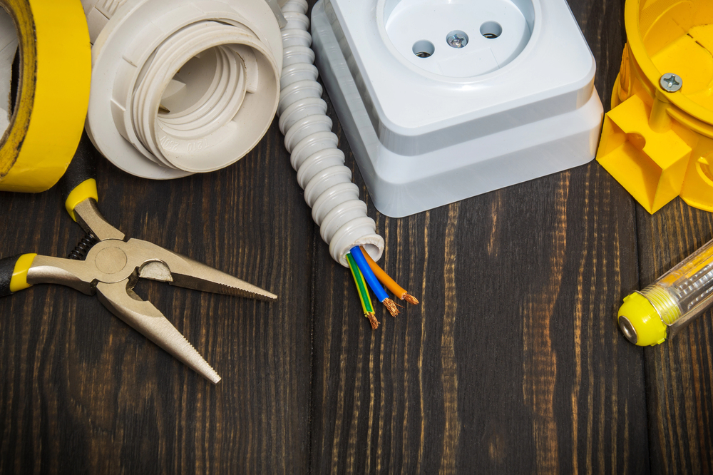 Assorted electrical tools and components for routine electrical repairs, including pliers, electrical tape, wires, an outlet, and a screwdriver, arranged on a wooden surface.