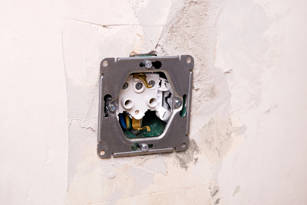 An exposed electrical outlet box with wiring, often seen during routine electrical repairs, is mounted on a wall with unfinished plaster.