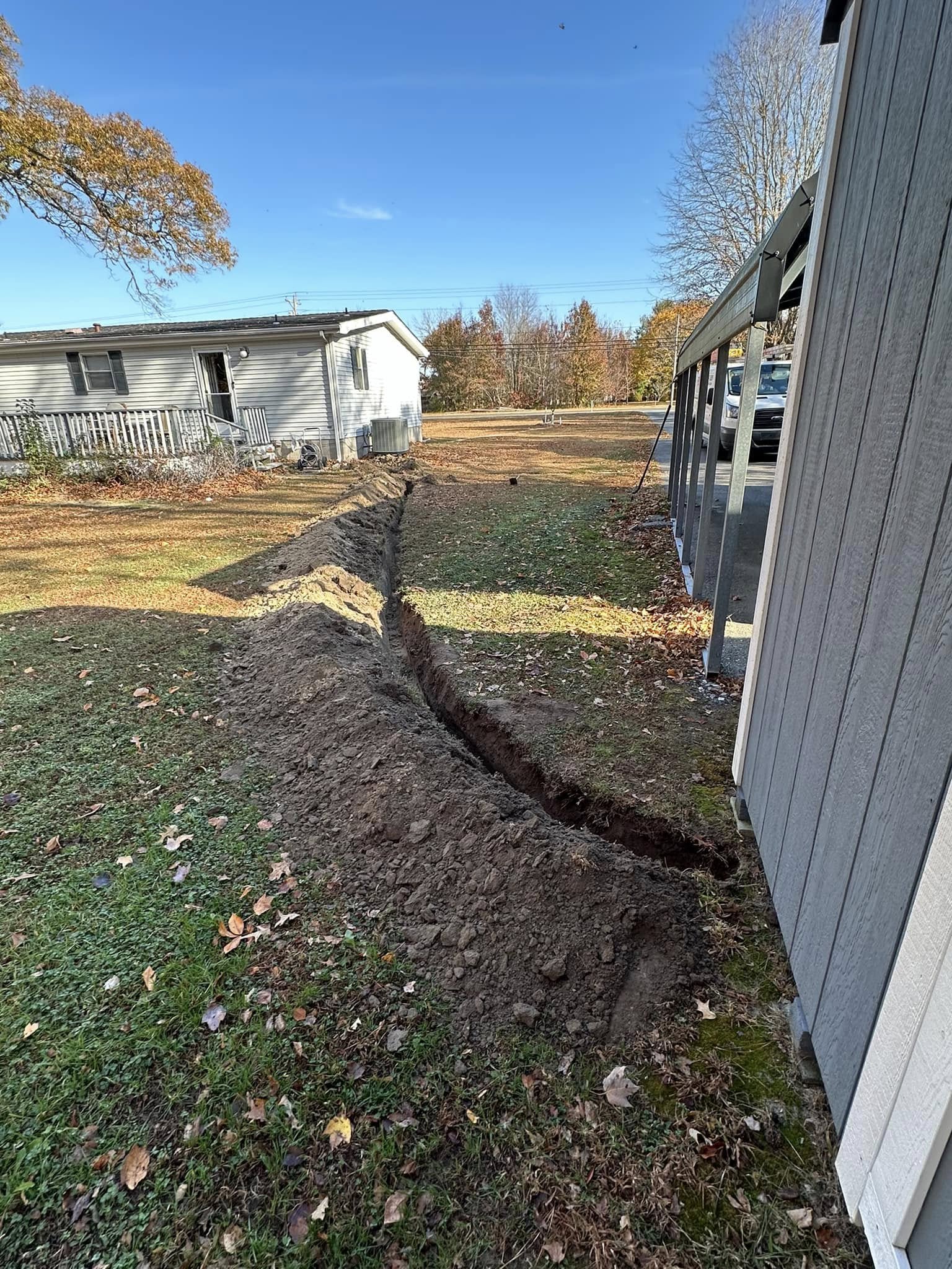 A freshly dug trench runs from a shed toward a house across a grassy yard on a clear day.